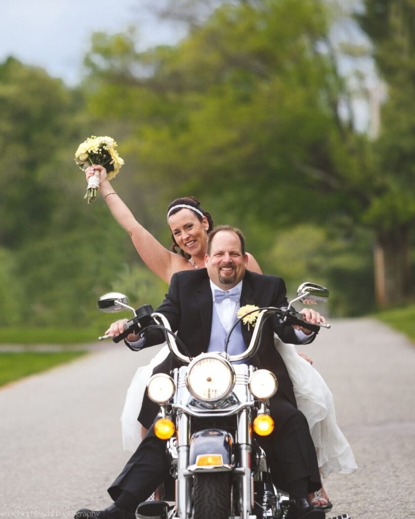 Smiling bride raising her yellow bouquet in the air while riding on the back of a motorcycle with her groom
